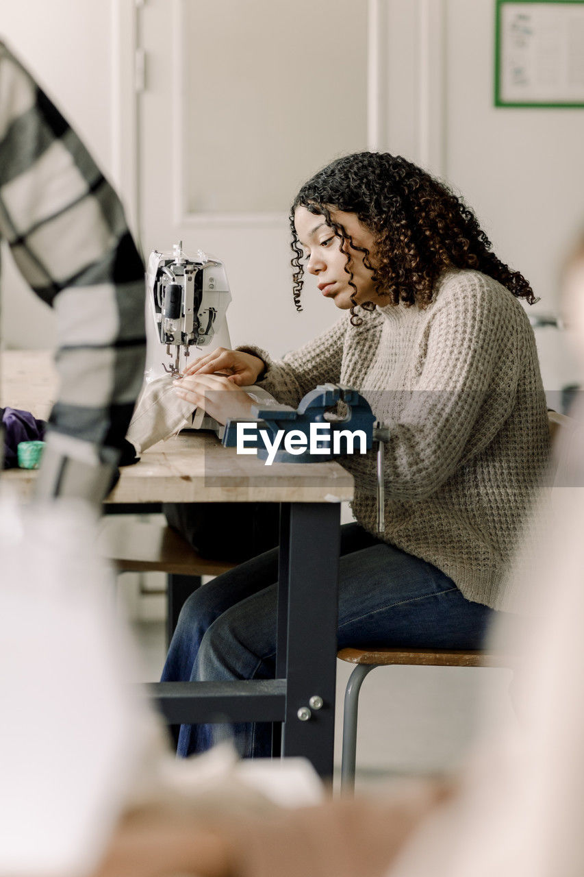 Teenage girl with curly hair using sewing machine in art class at high school