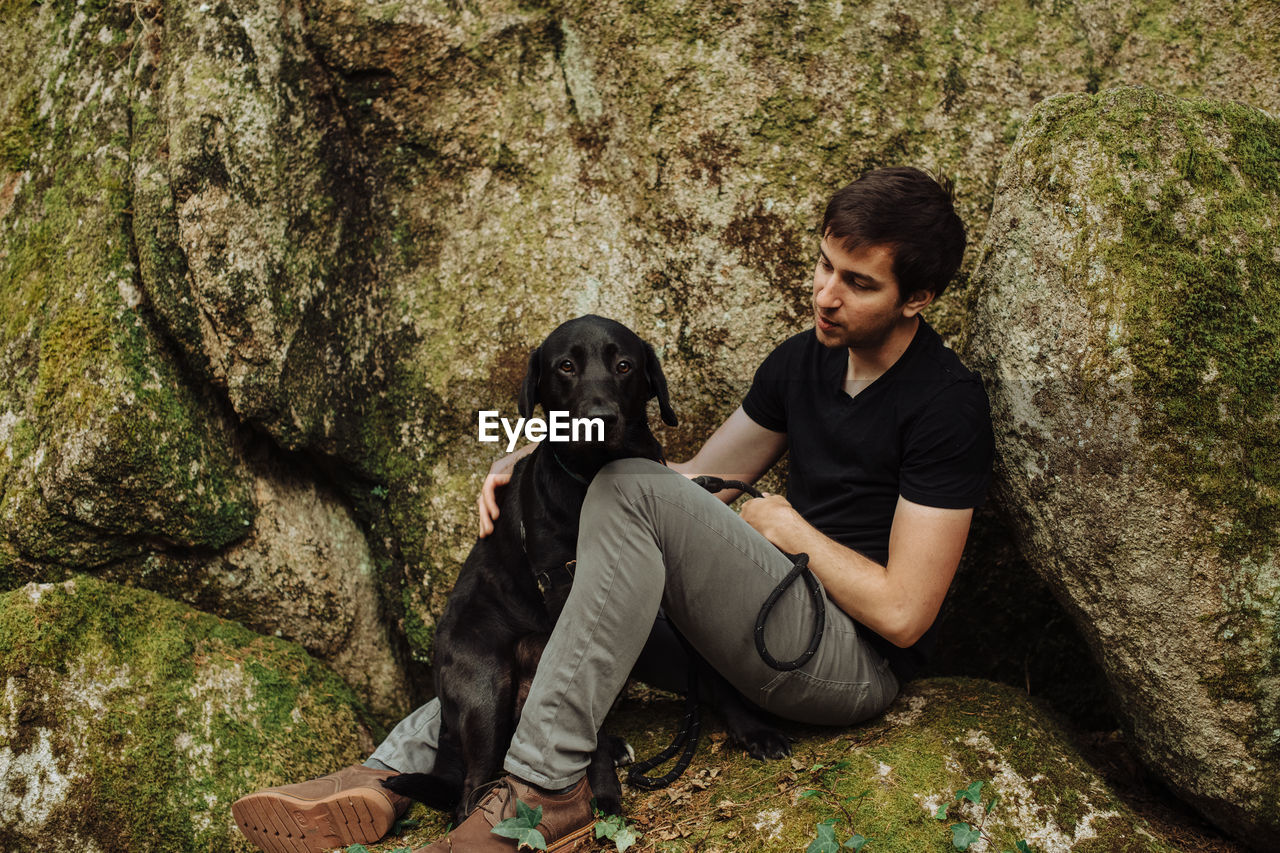 Young man with a black labrador retriever sitting on a mossy rock