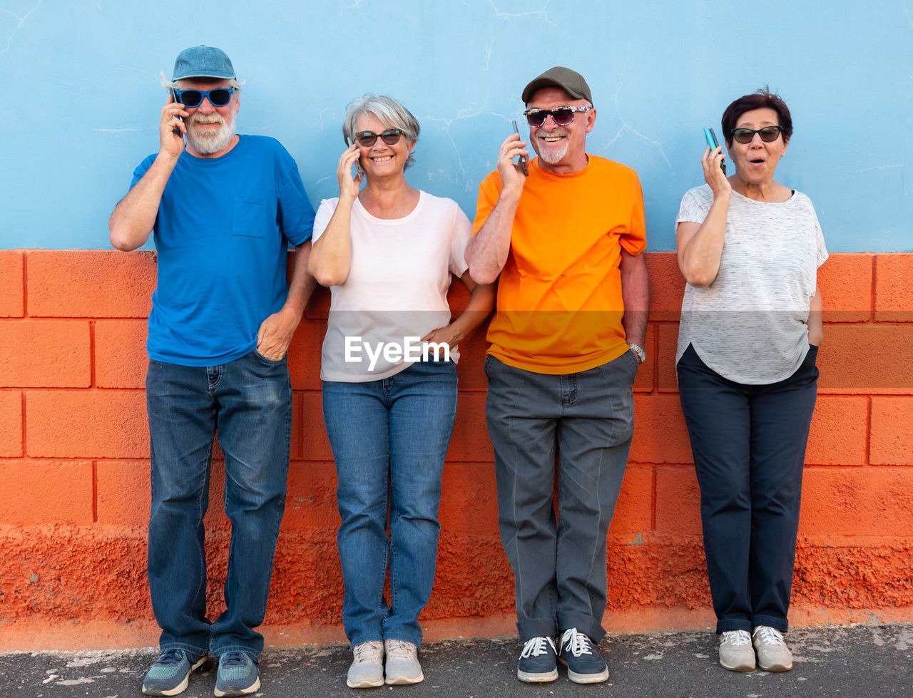 Full length of smiling senior couples talking on phone while standing against wall