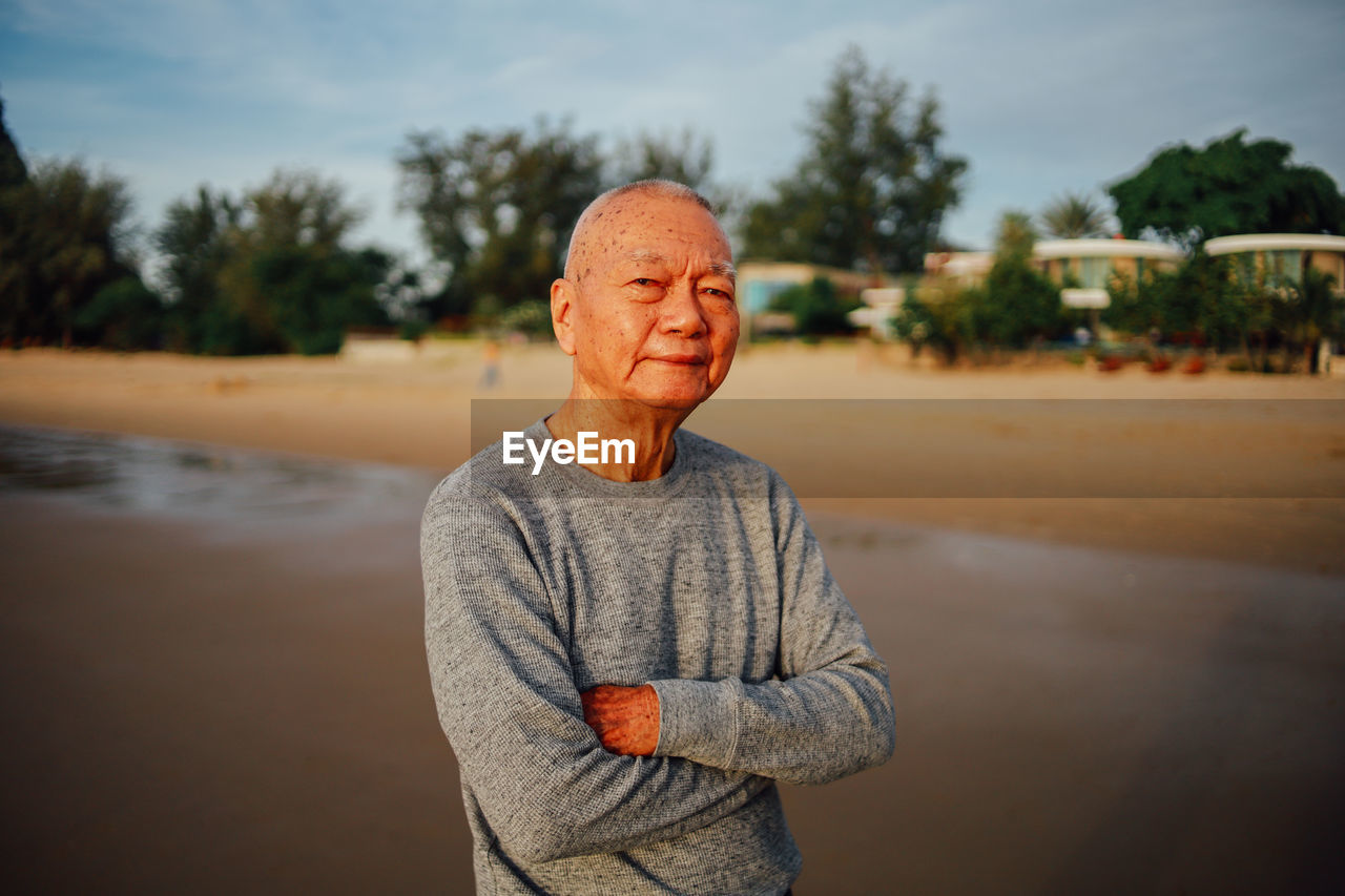 Portrait of senior man with arms crossed standing at beach against sky during sunset