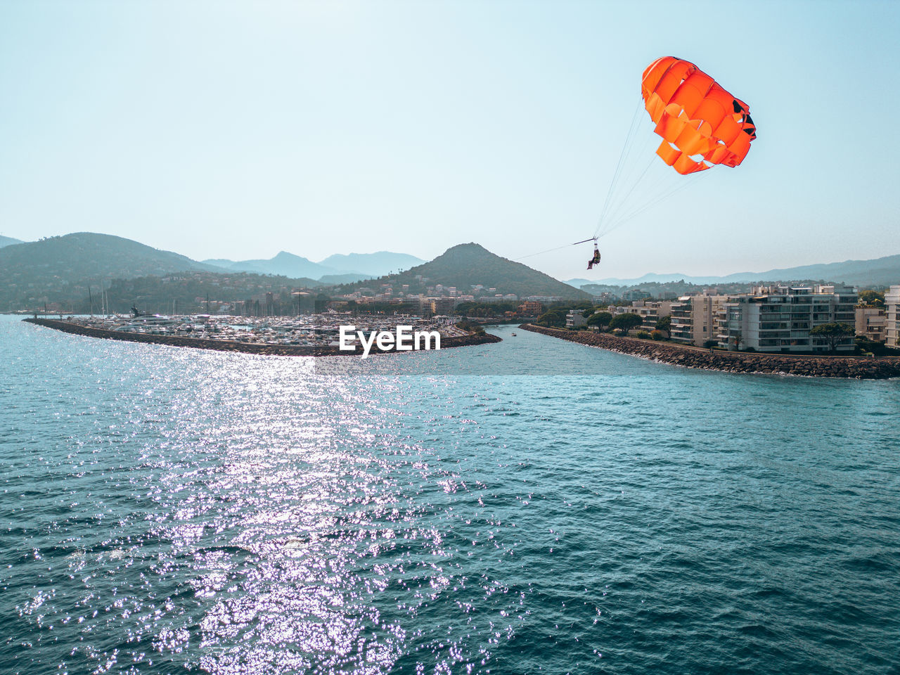 water, sailing, windsports, sky, nature, sea, sports, parasailing, transportation, adventure, mountain, mid-air, flying, day, travel destinations, scenics - nature, parachute, architecture, beauty in nature, blue, holiday, outdoors, travel, vacation, environment, water sports, motion, trip, clear sky, paragliding, kite sports, leisure activity, landscape, built structure, land, tranquility, sunny, city, extreme sports, rippled, tranquil scene, nautical vessel, mountain range, building exterior, no people