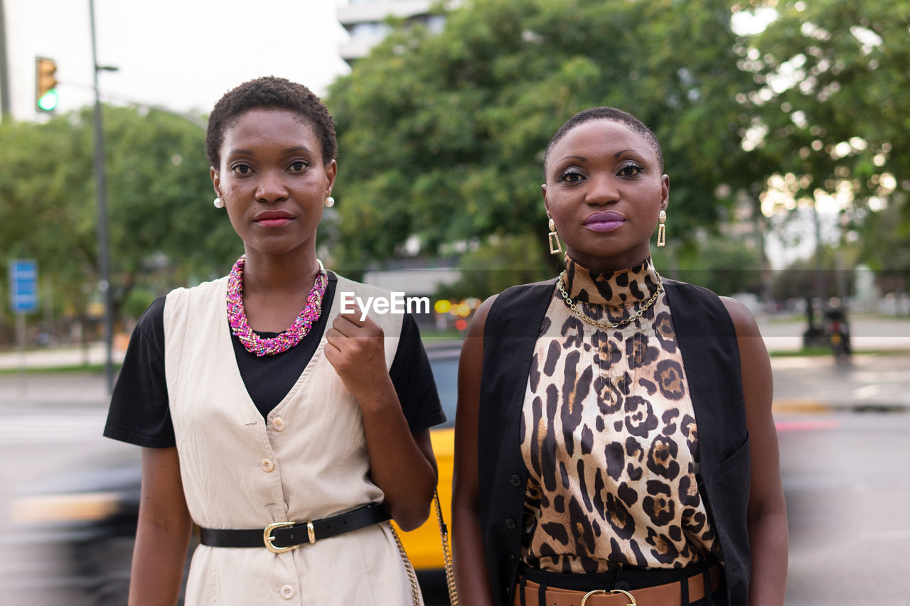 Feminine adult black women with short dark hair in fashionable outfits and accessories standing together on city street and looking at camera