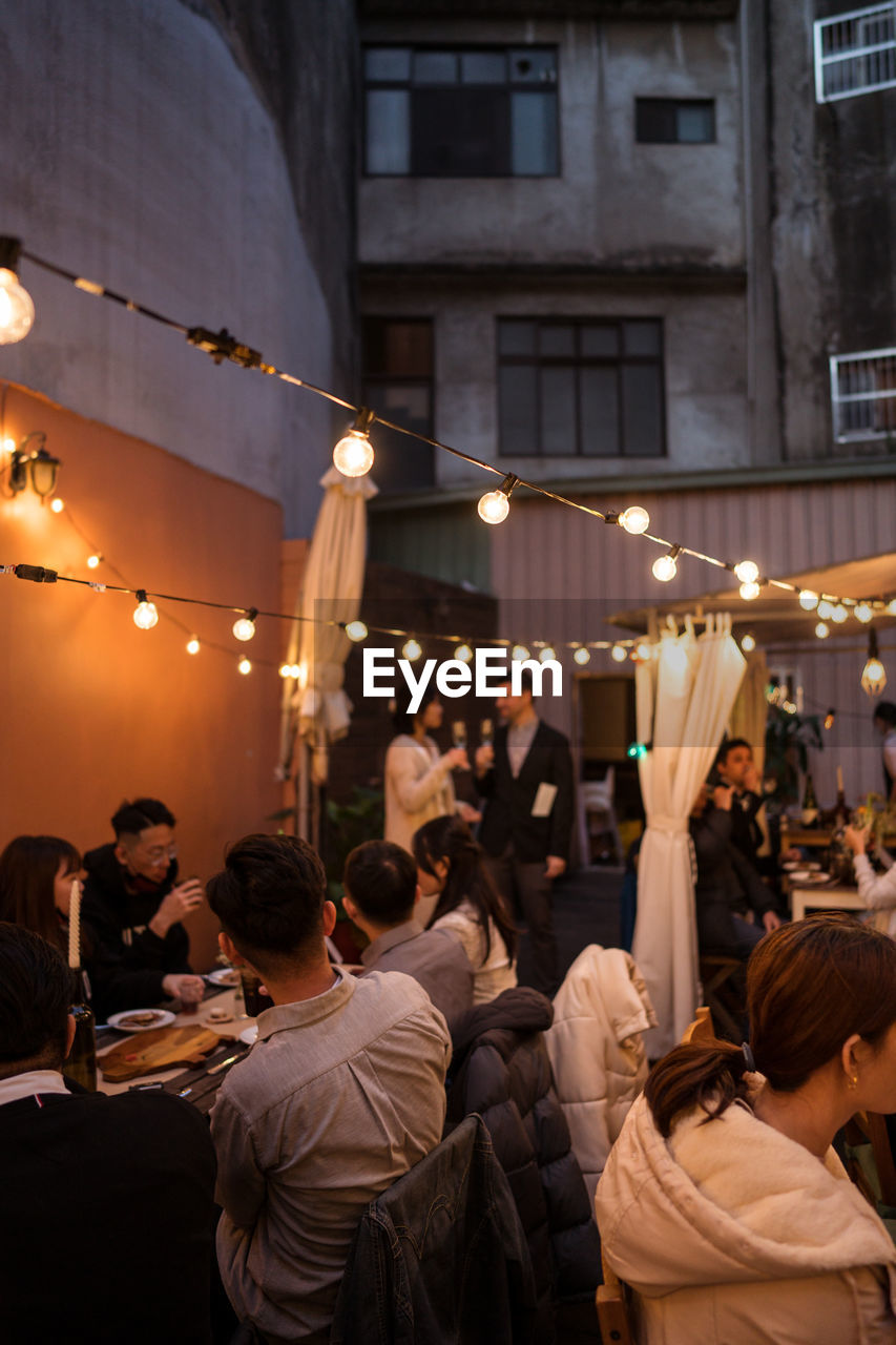 Group of guests celebrating wedding ceremony on street decorated with light bulb garlands and plants in dark evening