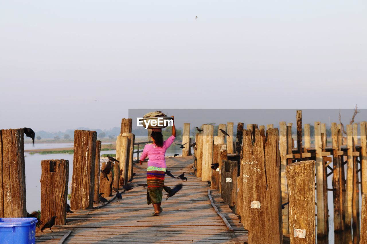 Woman with basket on head walking on pier over lake