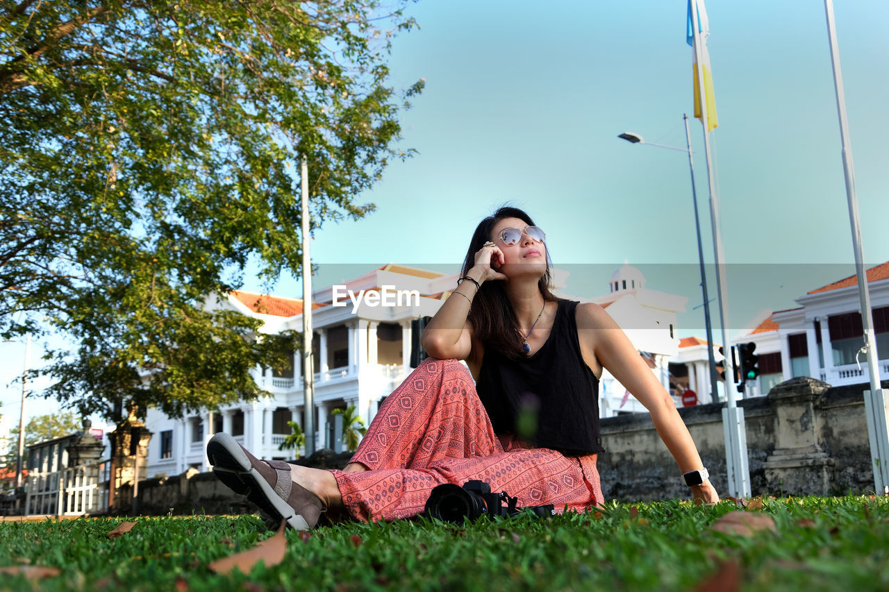 Smiling young woman sitting on grass against building and sky