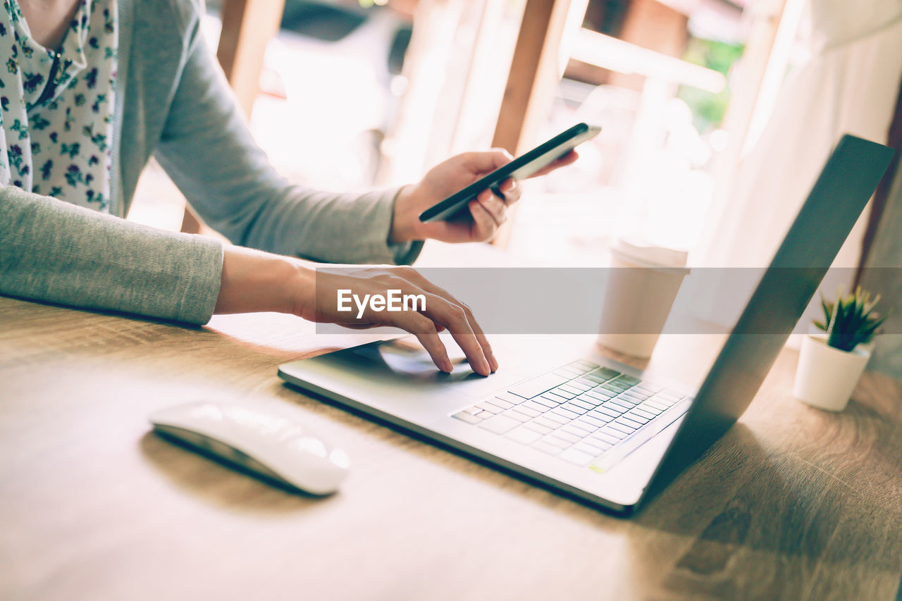 Midsection of woman using phone and laptop on table