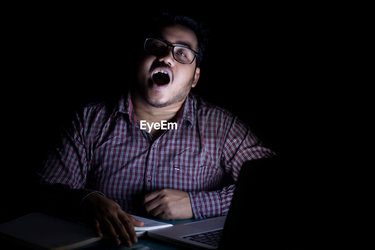 technology, one person, computer, laptop, using laptop, wireless technology, communication, adult, indoors, black background, glasses, men, eyeglasses, internet, copy space, studio shot, computer network, looking, young adult, sitting, person, dark, portrait, button down shirt, front view, business, casual clothing, mouth open, emotion, waist up, desk, working, table, computer equipment