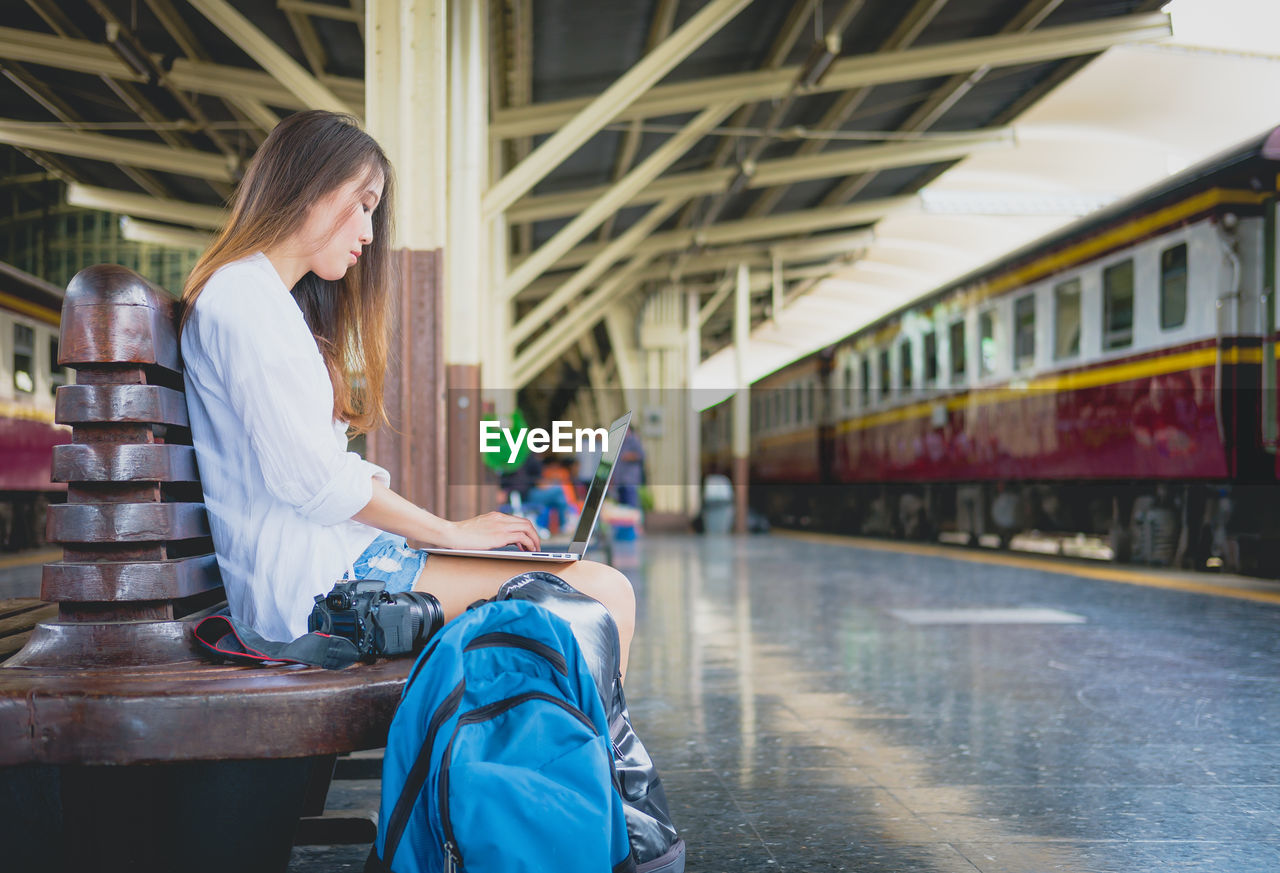 Side view of young woman using laptop while sitting on bench at railroad station