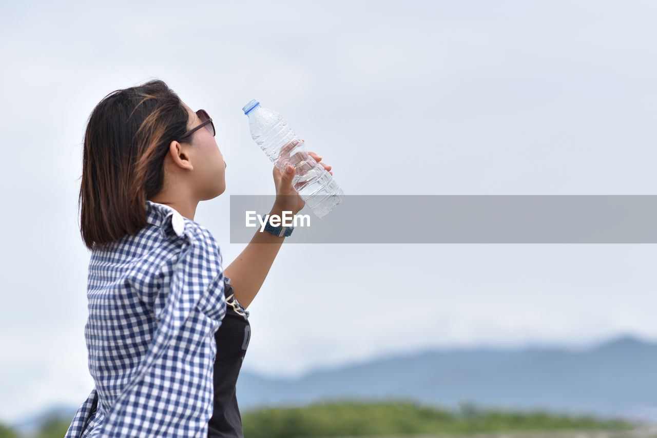 Side view of woman drinking water from bottle against sky