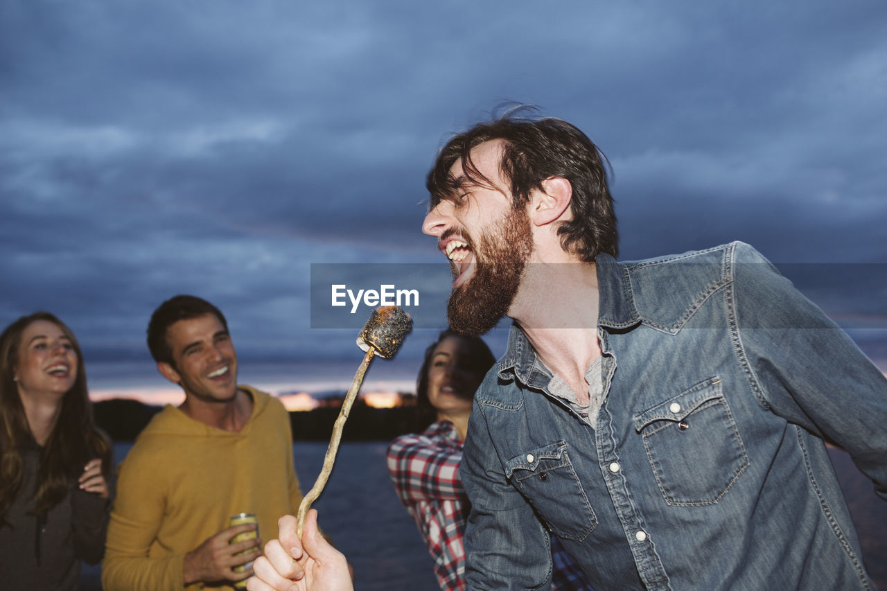 Smiling friends looking at man singing while holding roasted marshmallow on stick against cloudy sky