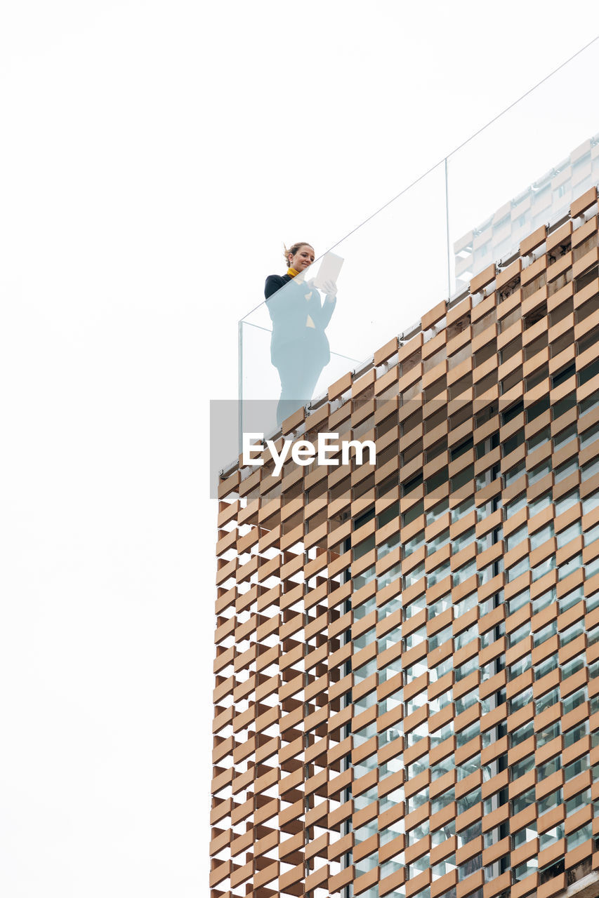 From below of female in stylish outfit standing on balcony of modern building with geometric elements on windows while using tablet near glass railings under bright sky