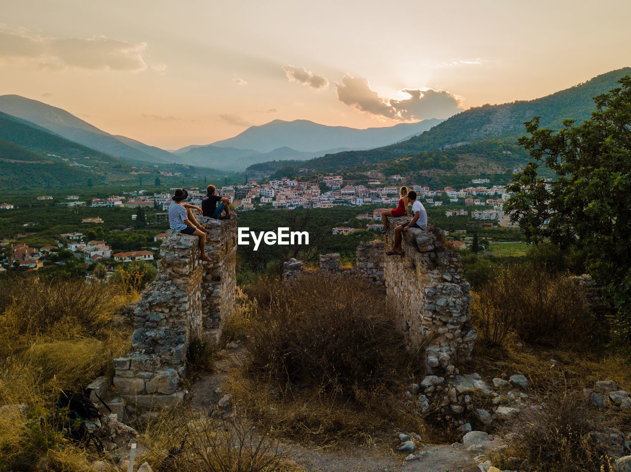 Friends sitting on old ruins against sky at sunset