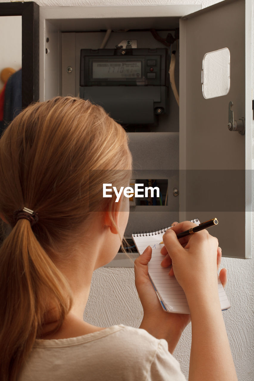 Rear view of woman writing in book while looking at fuse box