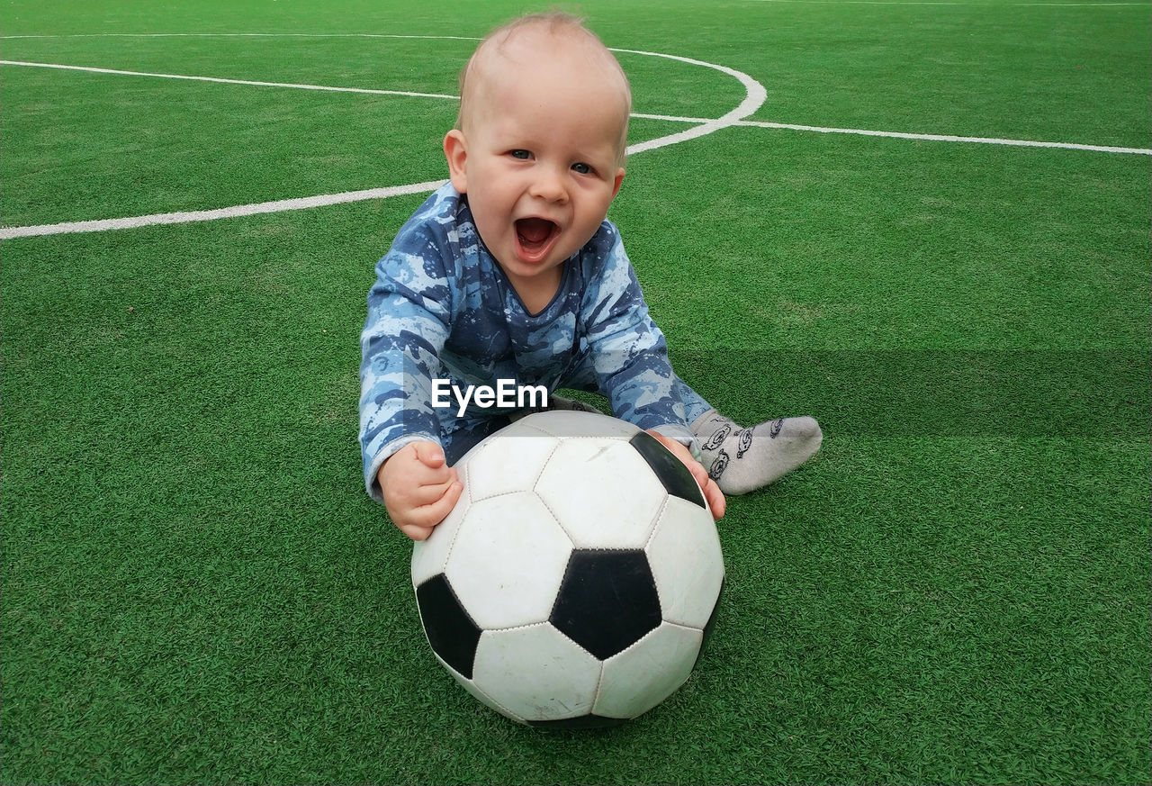 Portrait of cute baby boy with ball siting on soccer field