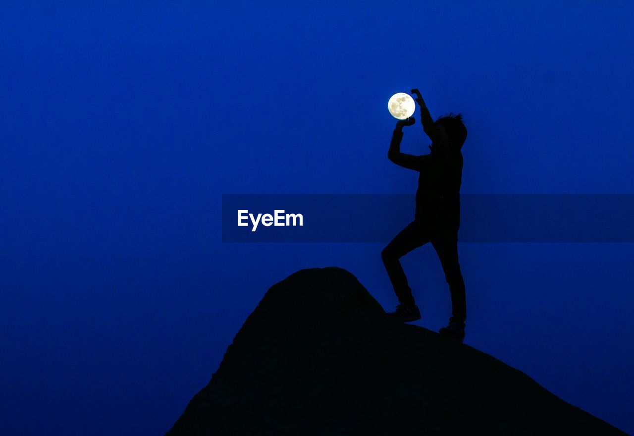 Optical illusion of silhouette man holding moon against blue sky at dusk