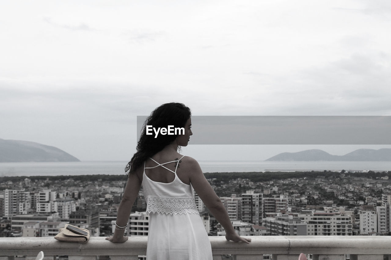 Rear view of woman standing by railing while looking at cityscape against sky