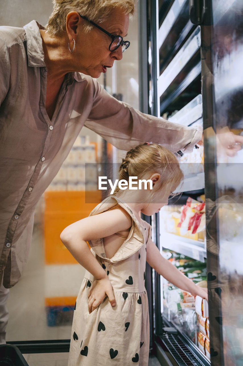 Side view of girl with hand on hip doing shopping from refrigerated section near grandmother at store
