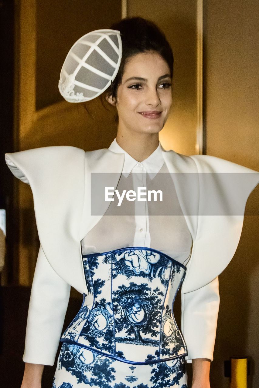 PORTRAIT OF YOUNG WOMAN STANDING AGAINST INDOORS