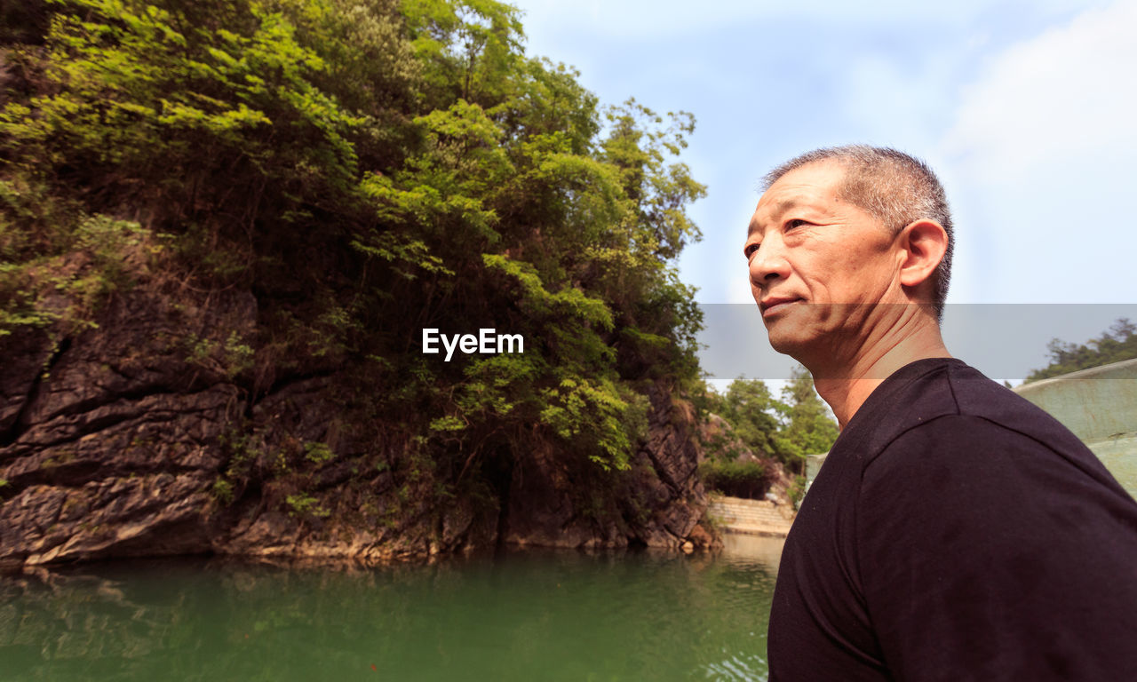 Thoughtful mature man looking away while standing at lakeshore against sky