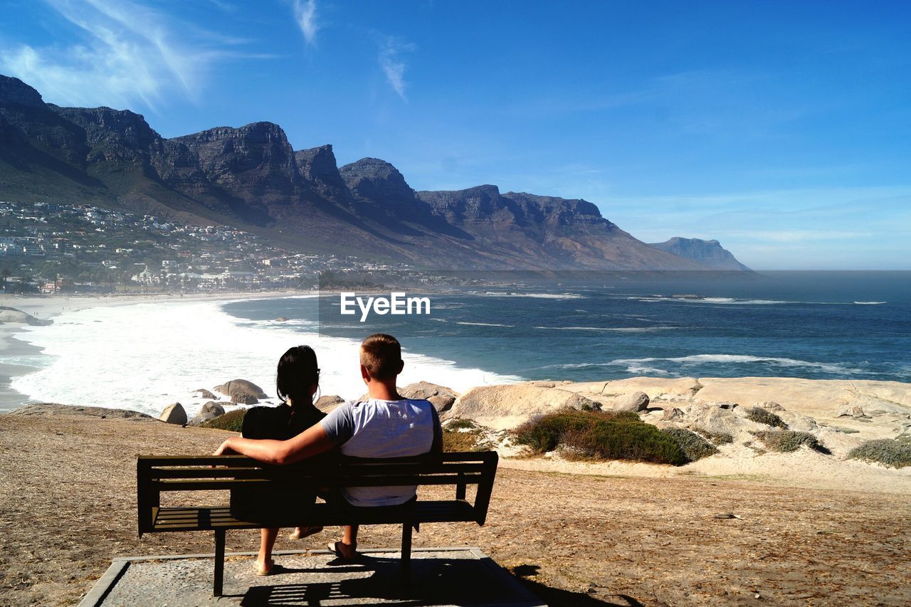 Rear view of couple sitting on bench by sea against sky