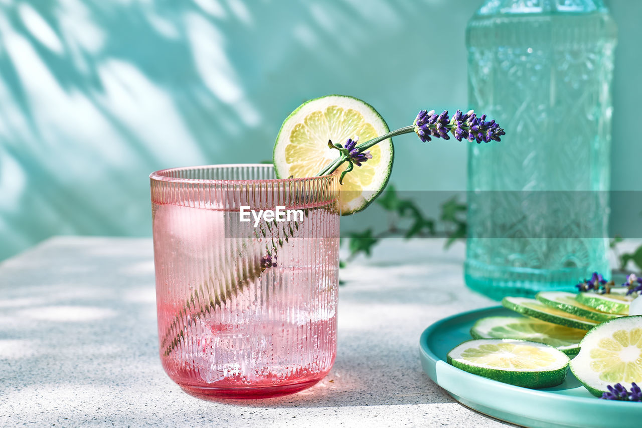Lavender lemonade with lime slices and lavender flower in pink glass near pastel light blue surface. 