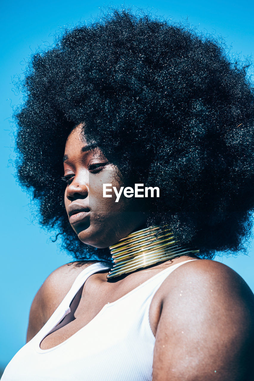 Young black woman in authentic golden necklace and afro hairstyle looking away on blurred background of countryside