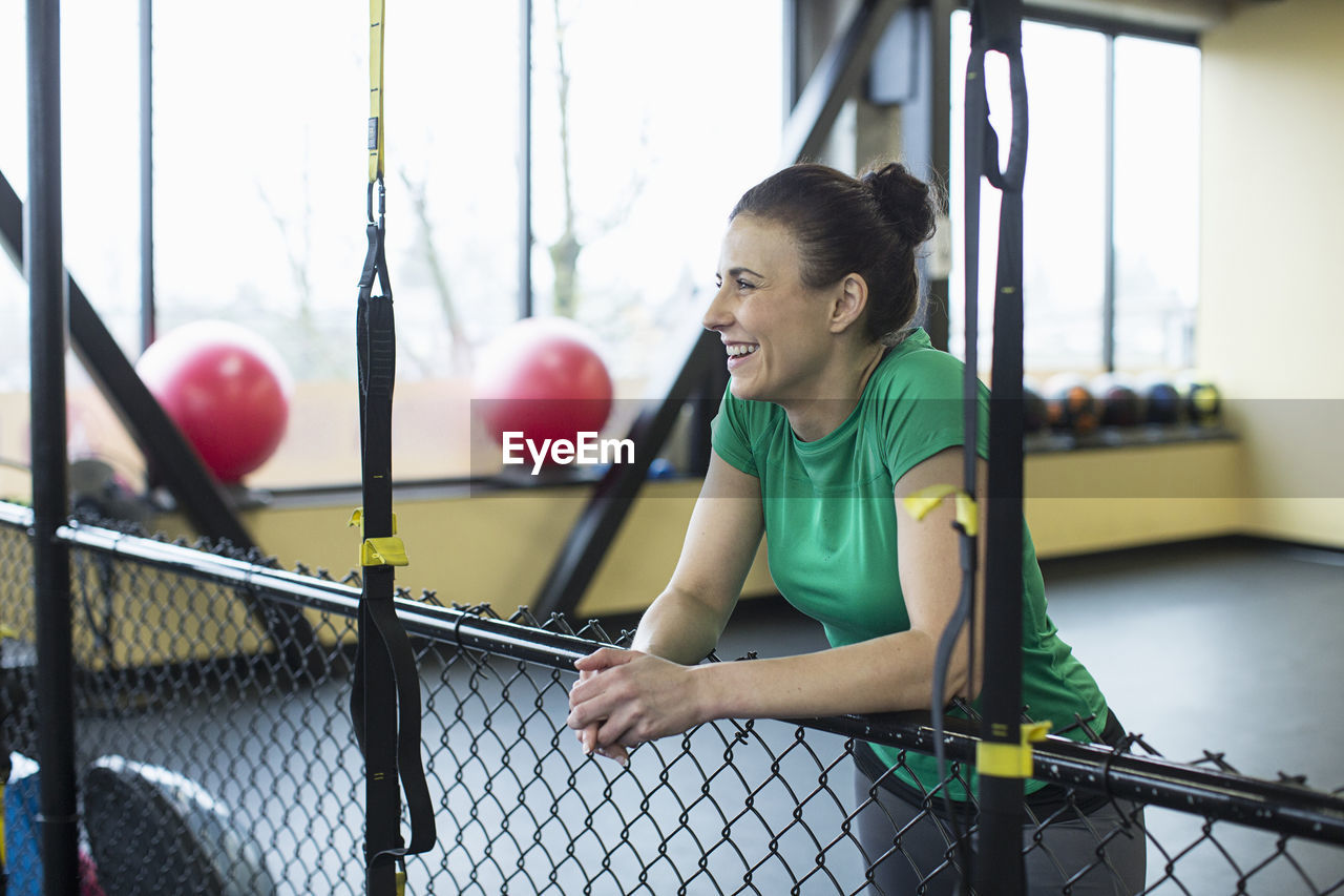 Female instructor smiling and looking away while leaning on chainlink fence in gym