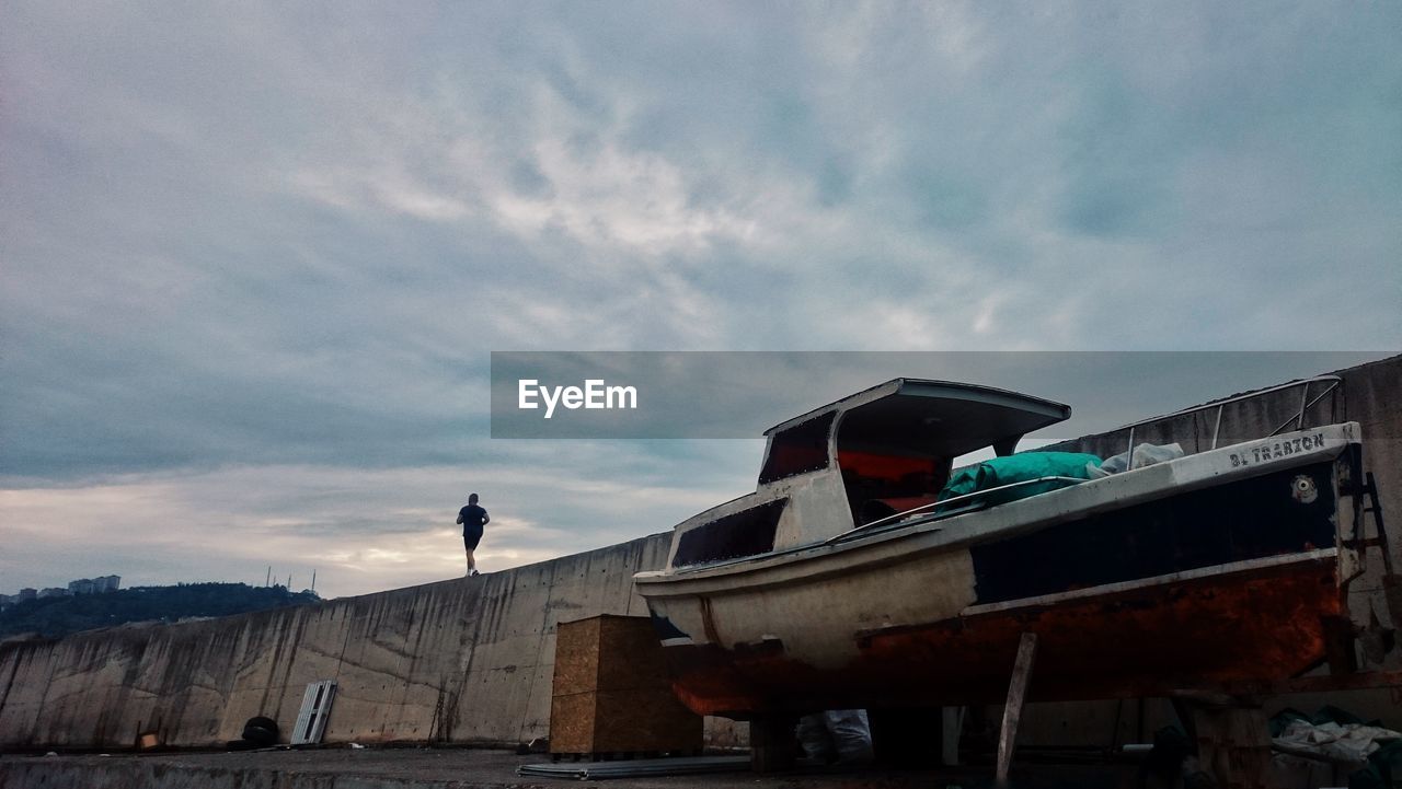 Low angle view of man running on retaining wall by boat moored on pier against cloudy sky