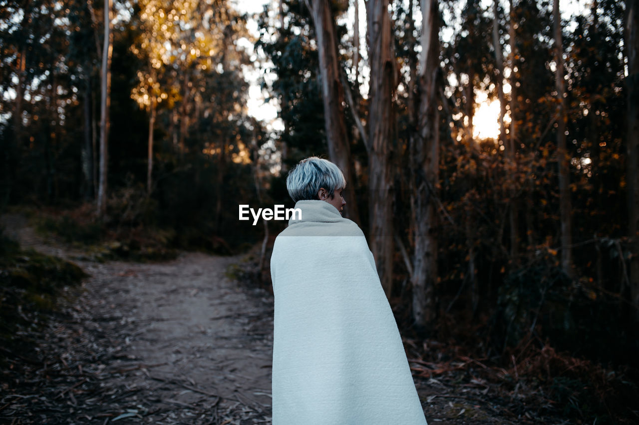 Back view of anonymous female with short hair covered with white blanket strolling on rural path in woods on evening time