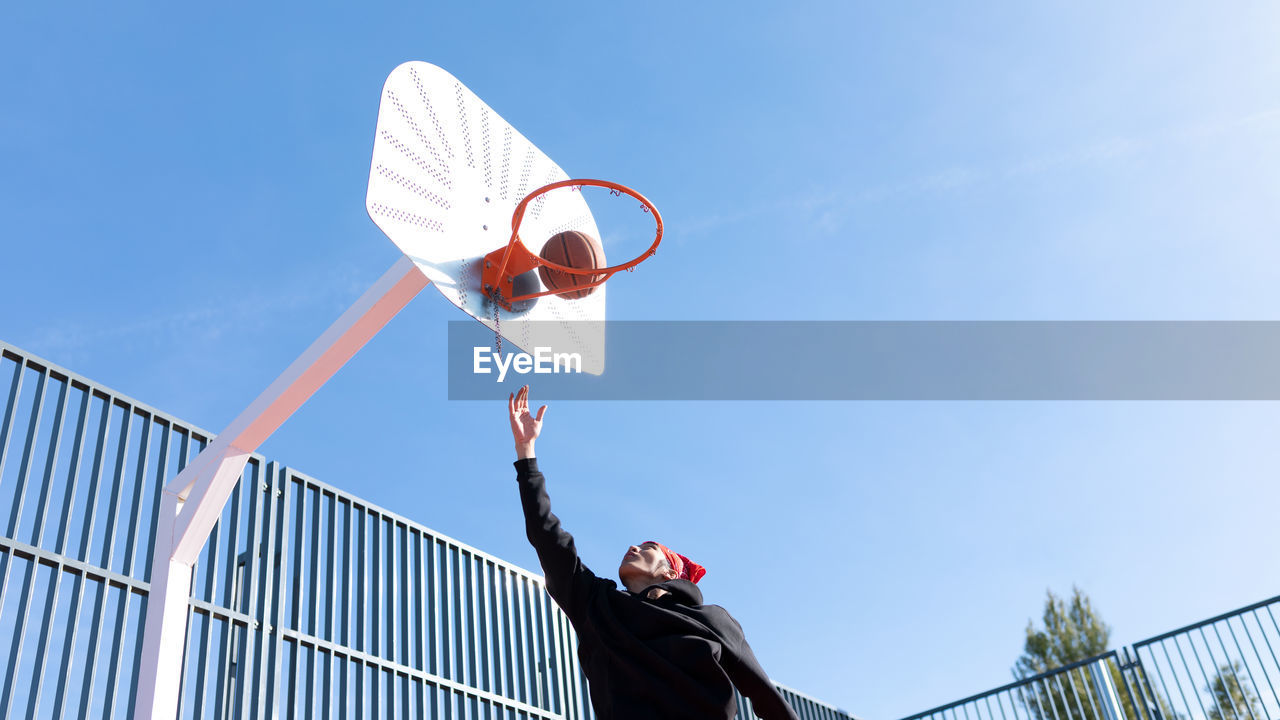 Active man throwing ball into basketball hoop with raised arm while playing on sports ground on sunny day during training