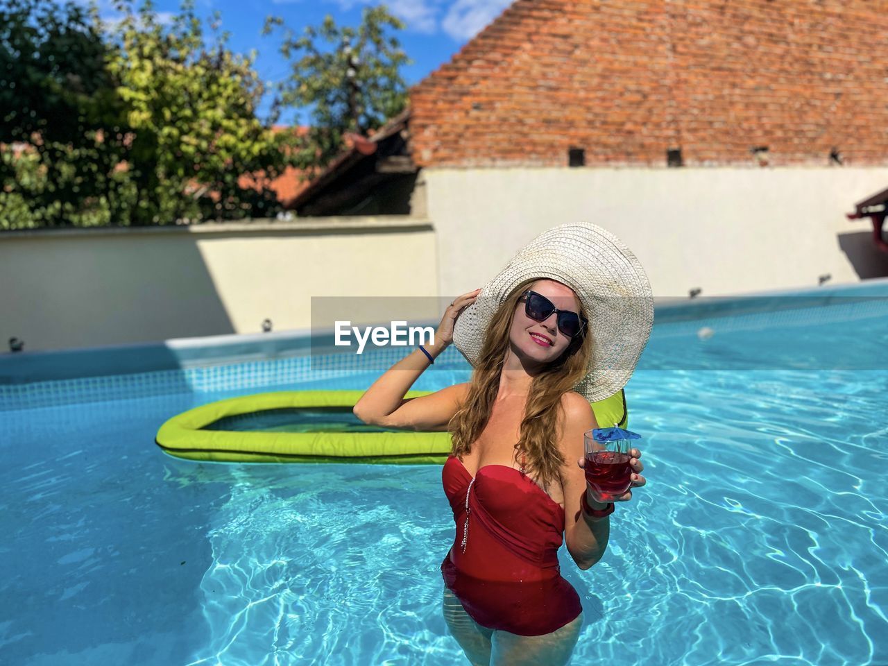 Young fashionable woman wearing red swimming suit and white hat holding a drink in the pool