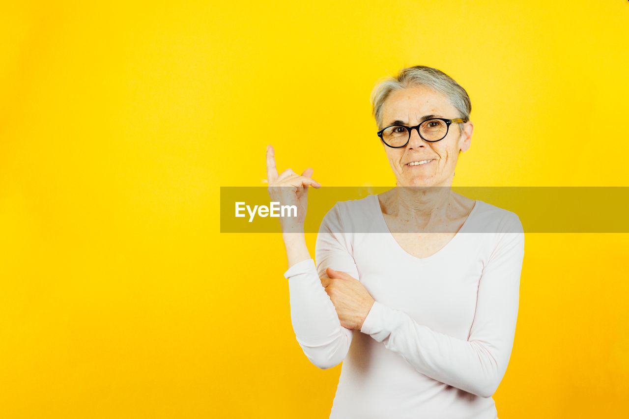 yellow, eyeglasses, glasses, adult, one person, indoors, waist up, portrait, studio shot, person, colored background, smiling, yellow background, copy space, clothing, front view, mature adult, looking at camera, emotion, finger, happiness, casual clothing, human face, standing, wall - building feature, women, business, hand, senior adult, men