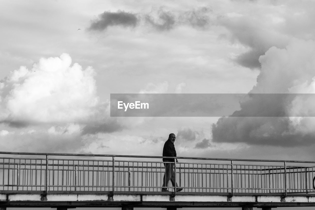 Side view of man walking on bridge against cloudy sky