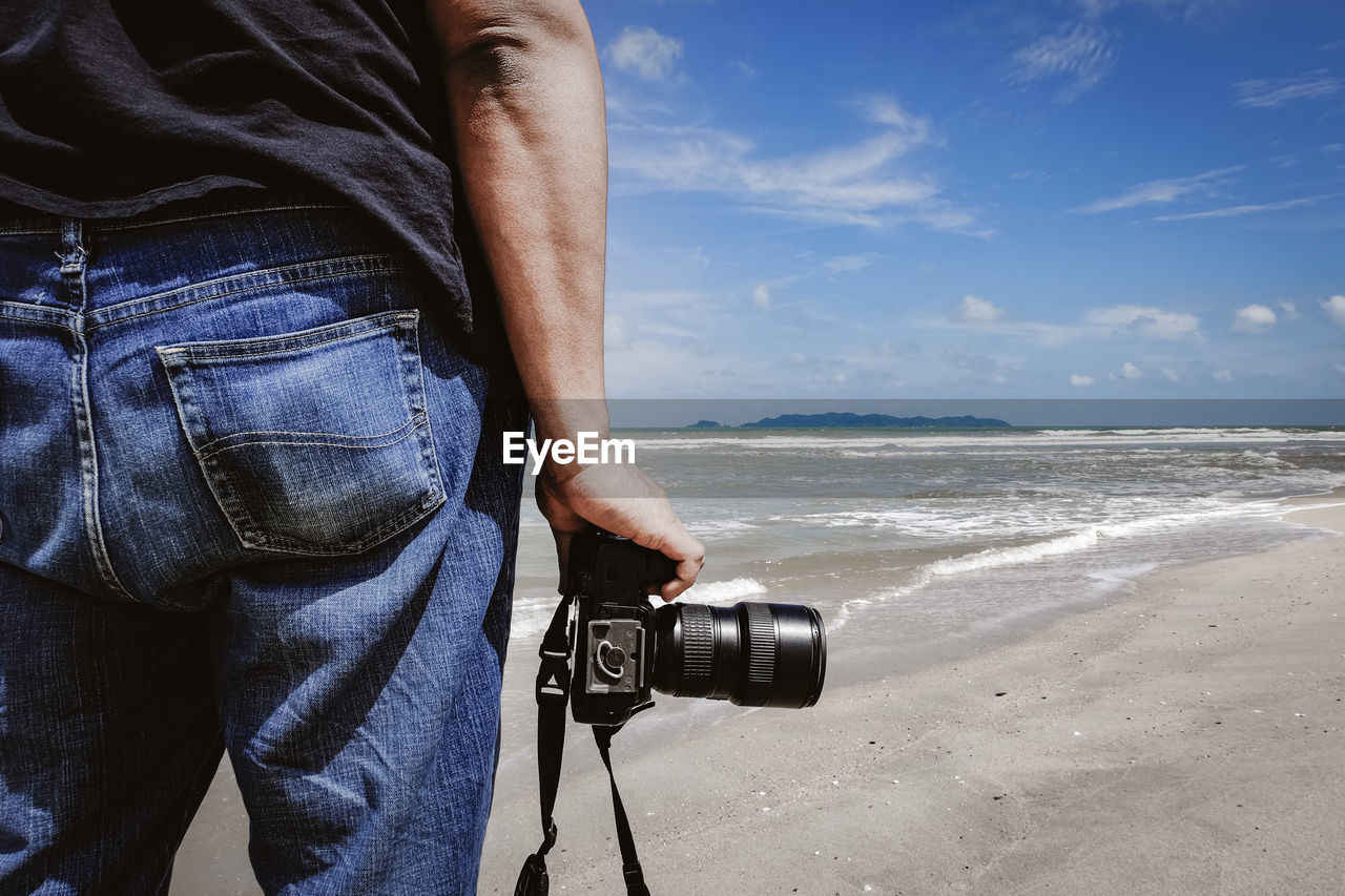 Midsection of man with camera standing at beach against sky