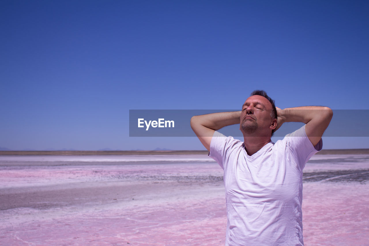 Mature man with eyes closed standing at beach against clear blue sky