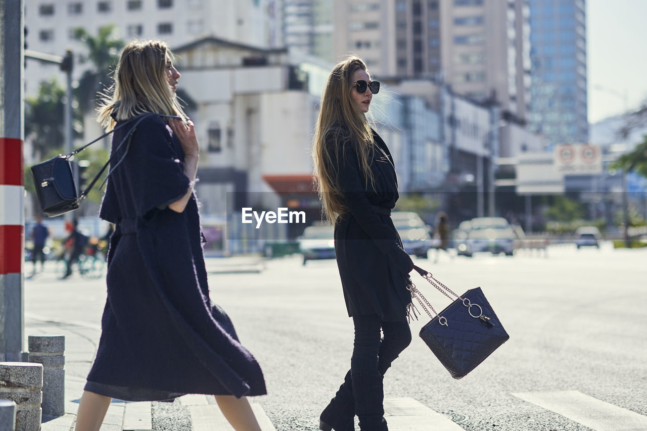 Side view of blonde stylish young female partners in trendy apparel strolling on asphalt road in city