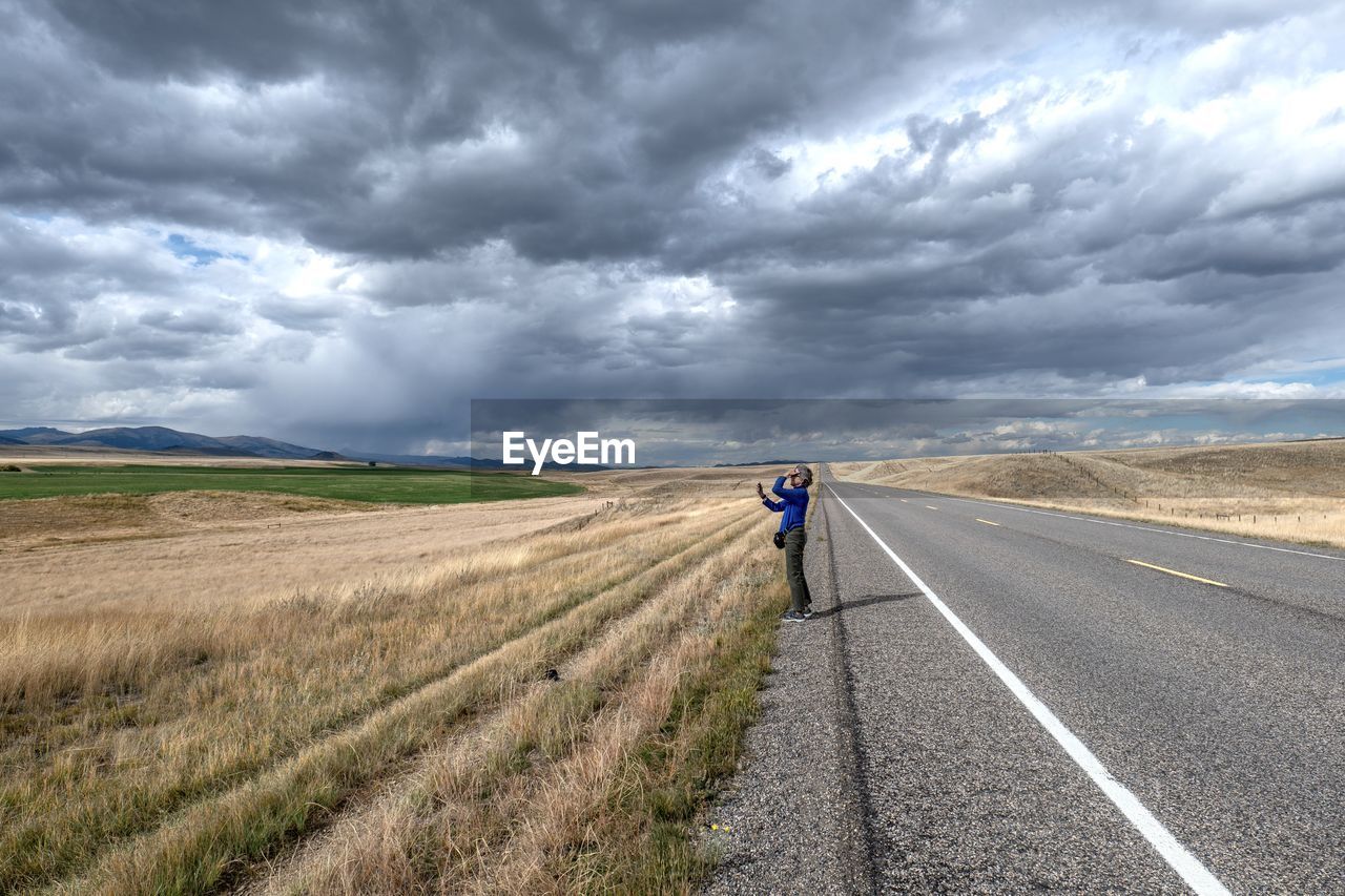 cloud, landscape, road, sky, environment, transportation, horizon, land, nature, highway, rural scene, scenics - nature, field, storm, prairie, beauty in nature, dramatic sky, plain, one person, storm cloud, overcast, grass, rural area, cloudscape, adult, travel, the way forward, horizon over land, motion, plant, non-urban scene, outdoors, day, remote, grassland, sign, journey, full length, asphalt, moody sky, solitude, country road, agriculture, vanishing point, infrastructure, diminishing perspective