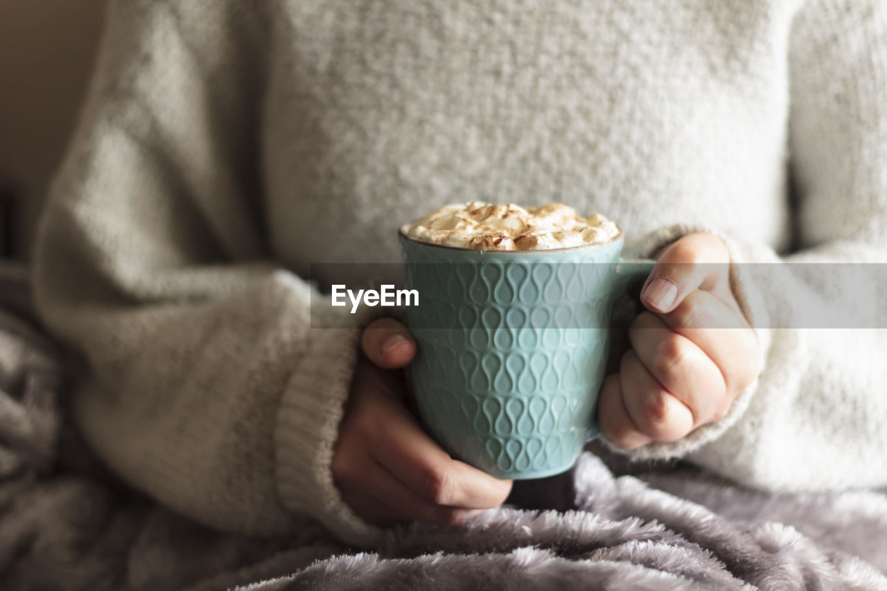 Woman covered with blanket holding mug of hot drink with whipped cream