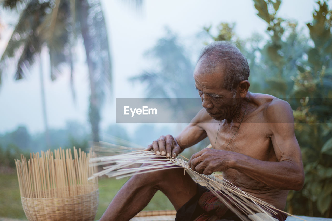 Shirtless man weaving wicker basket against trees