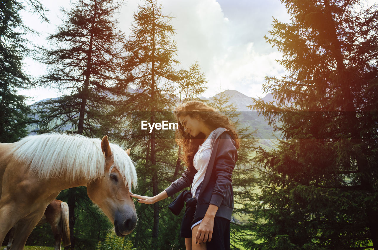 Woman touching horse while standing against trees