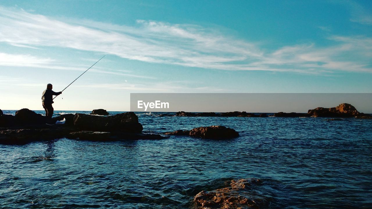 Man fishing in sea against cloudy sky