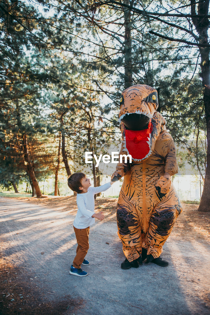 Boy with person wearing dinosaur costume standing on road in park