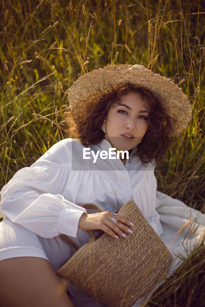 Portrait of a curly-haired woman, wicker hat and with a basket lying on a field with grass in autumn