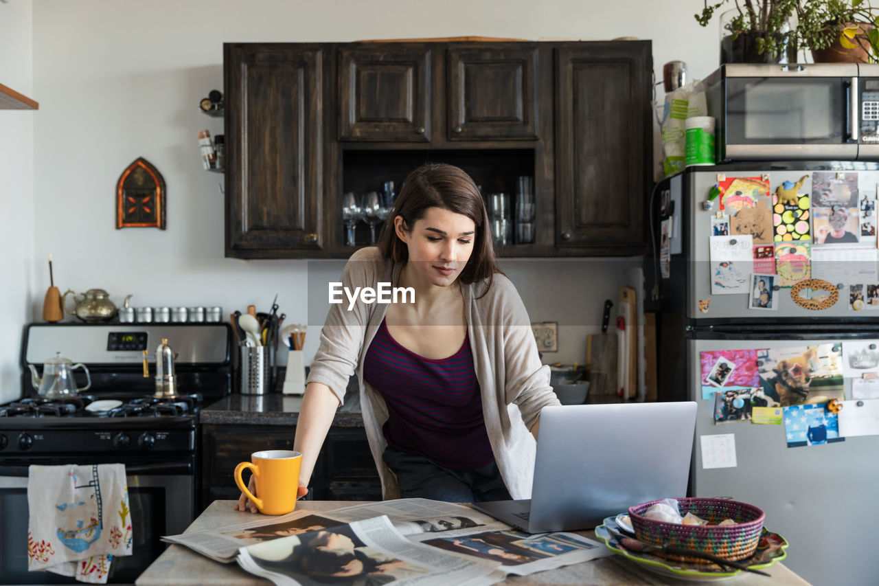 Young woman using laptop while having coffee at table in kitchen