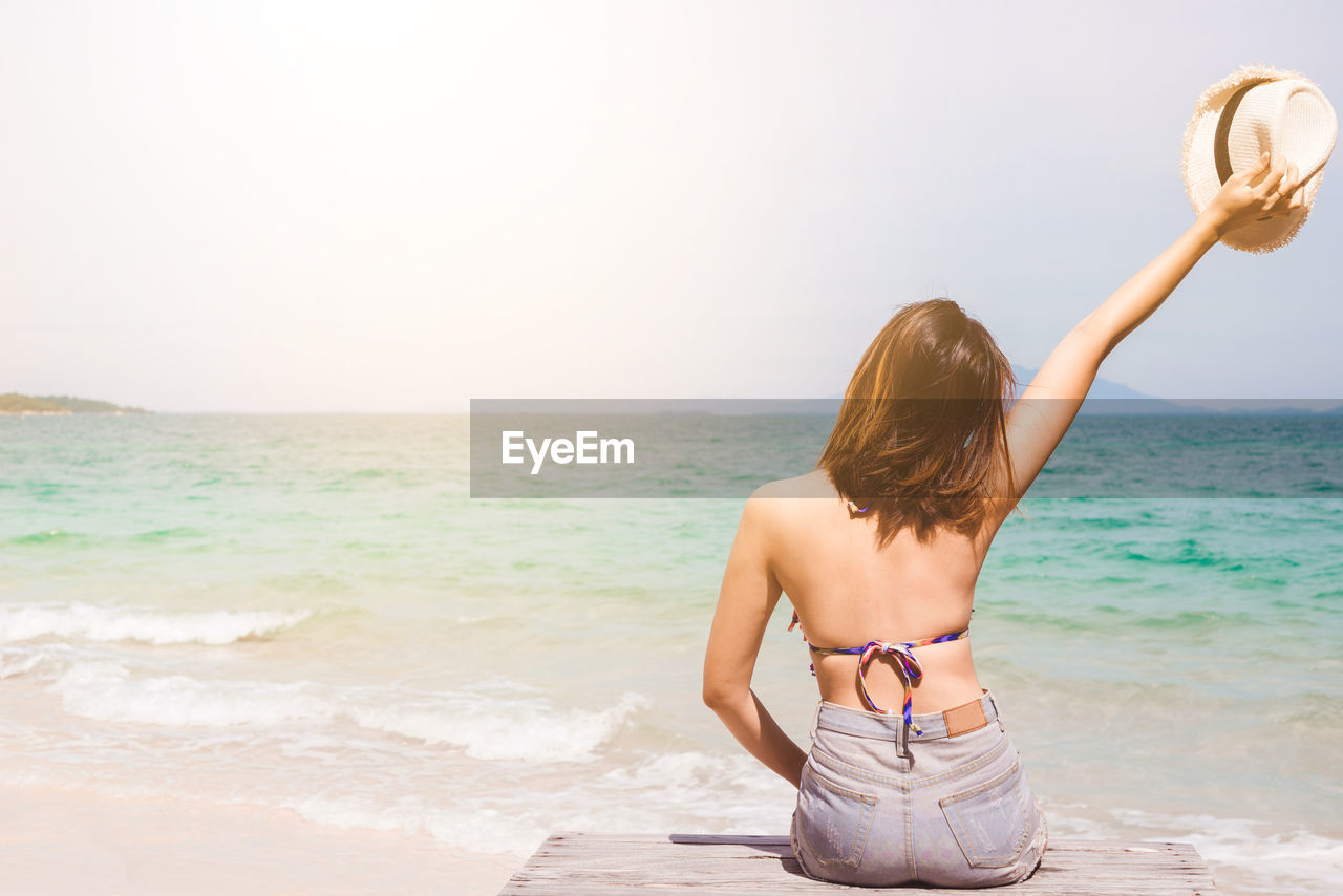 Rear view of woman holding hat while sitting by sea during sunny day