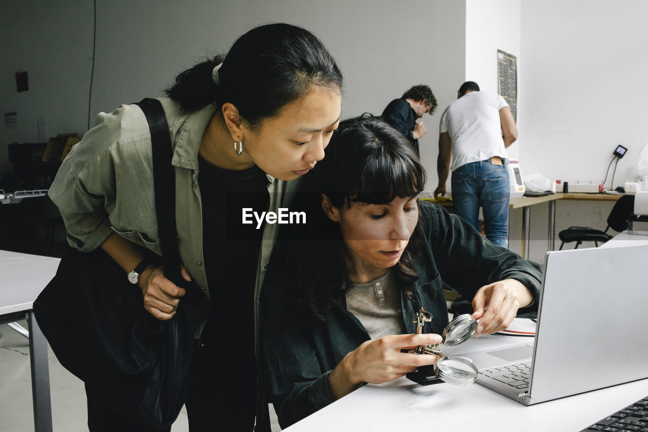Customer standing near focused female technician examining laptop through magnifying glass at repair shop