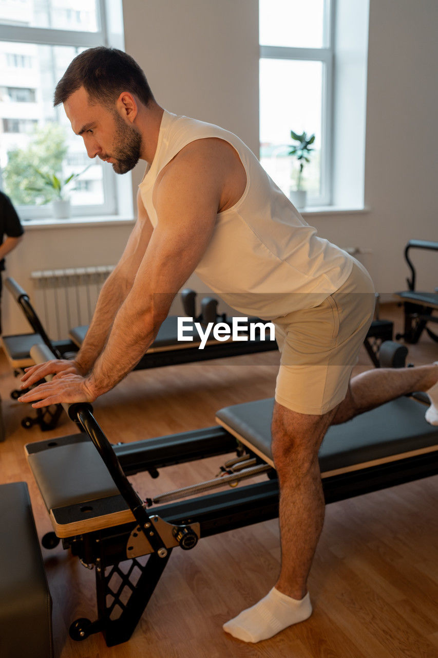 adult, indoors, lifestyles, arm, room, exercising, physical fitness, men, sport venue, limb, wellbeing, sports, young adult, muscular build, side view, human leg, fitness professional, sports training, person, health club, sports clothing, one person, strength, gym, full length, concentration, vitality, exercise equipment, activity, body conscious, clothing, sitting, window, equipment