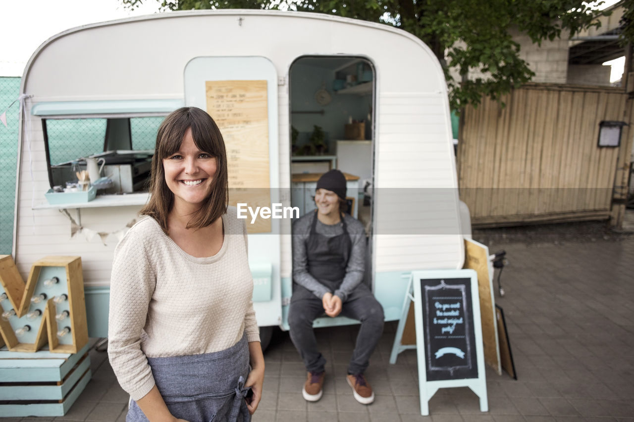 Portrait of happy female owner standing on street while coworker sitting in food truck