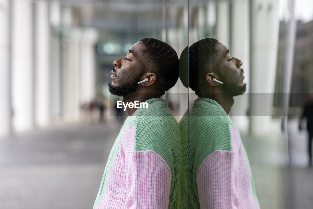 Man with eyes closed listening music through wireless in-ear headphones leaning on glass wall