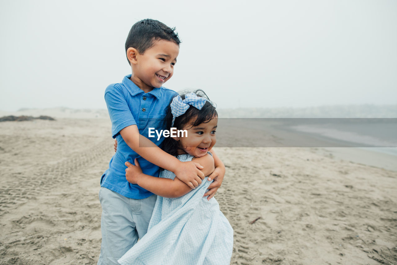 Young siblings smile and embrace at beach looking toward the waves